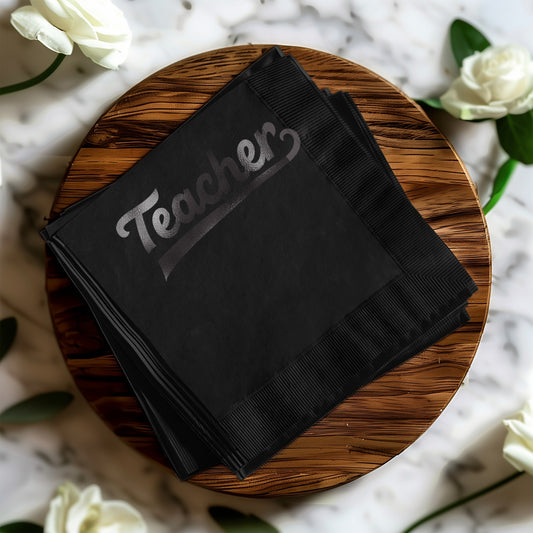 Black cocktail napkins with "Teacher" in silver lettering, stacked on a wooden board.